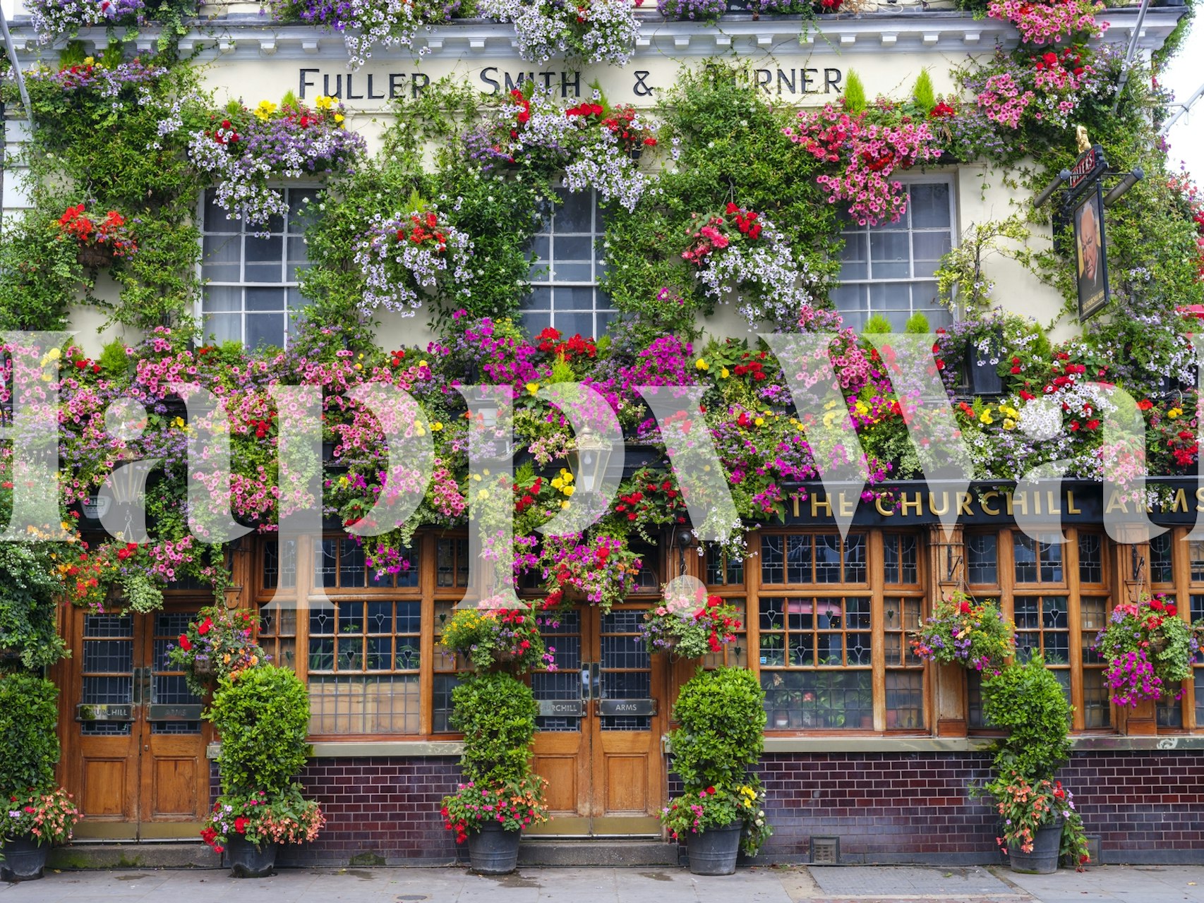 Το τοιχογραφία του The Churchill Arms pub με πλούσιες κρεμαστές γλάστρες λουλουδιών και κλασική βρετανική πρόσοψη