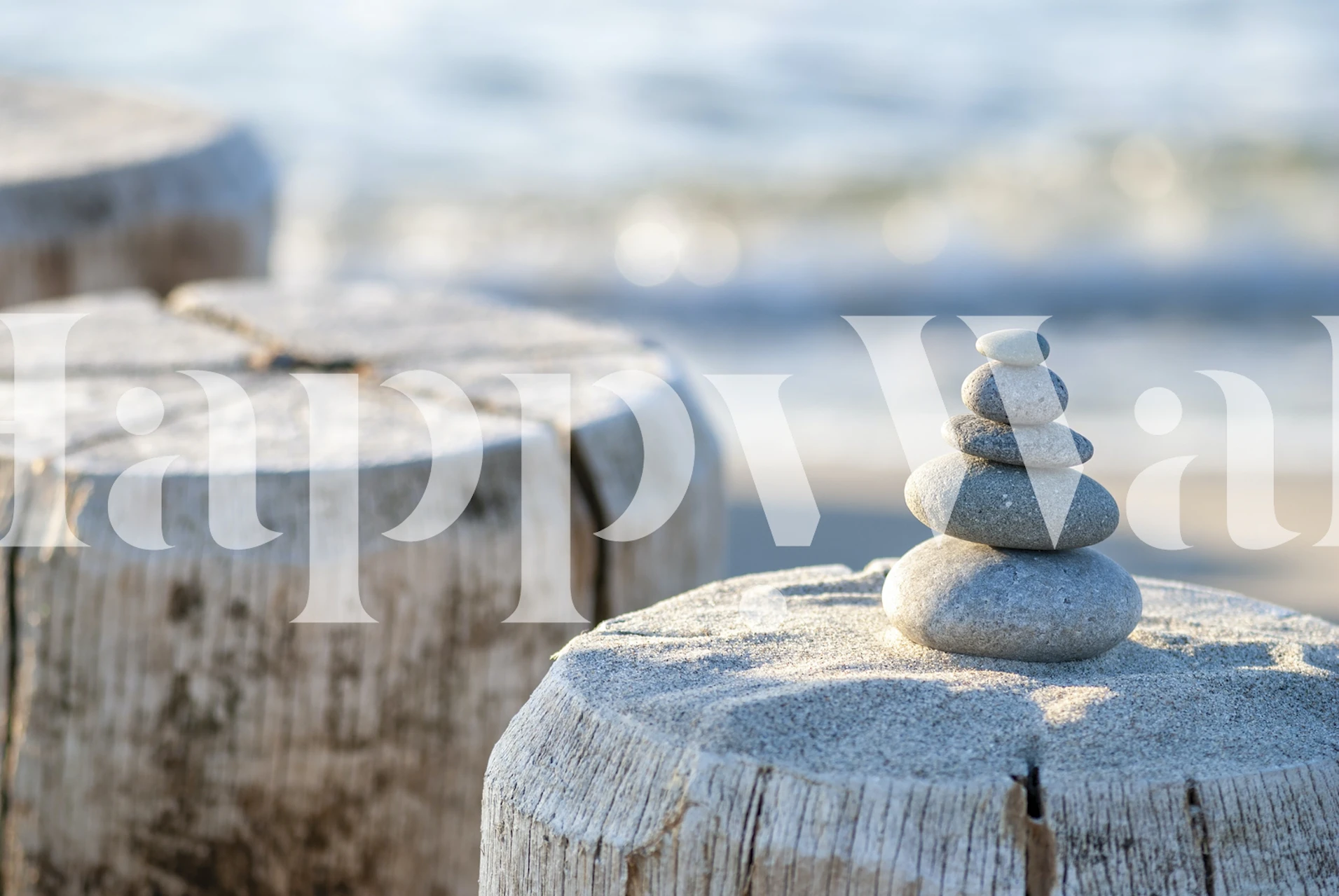 Pedras de seixo empilhadas sobre um toco de madeira com um fotomural vinílico de fundo de praia com foco suave.