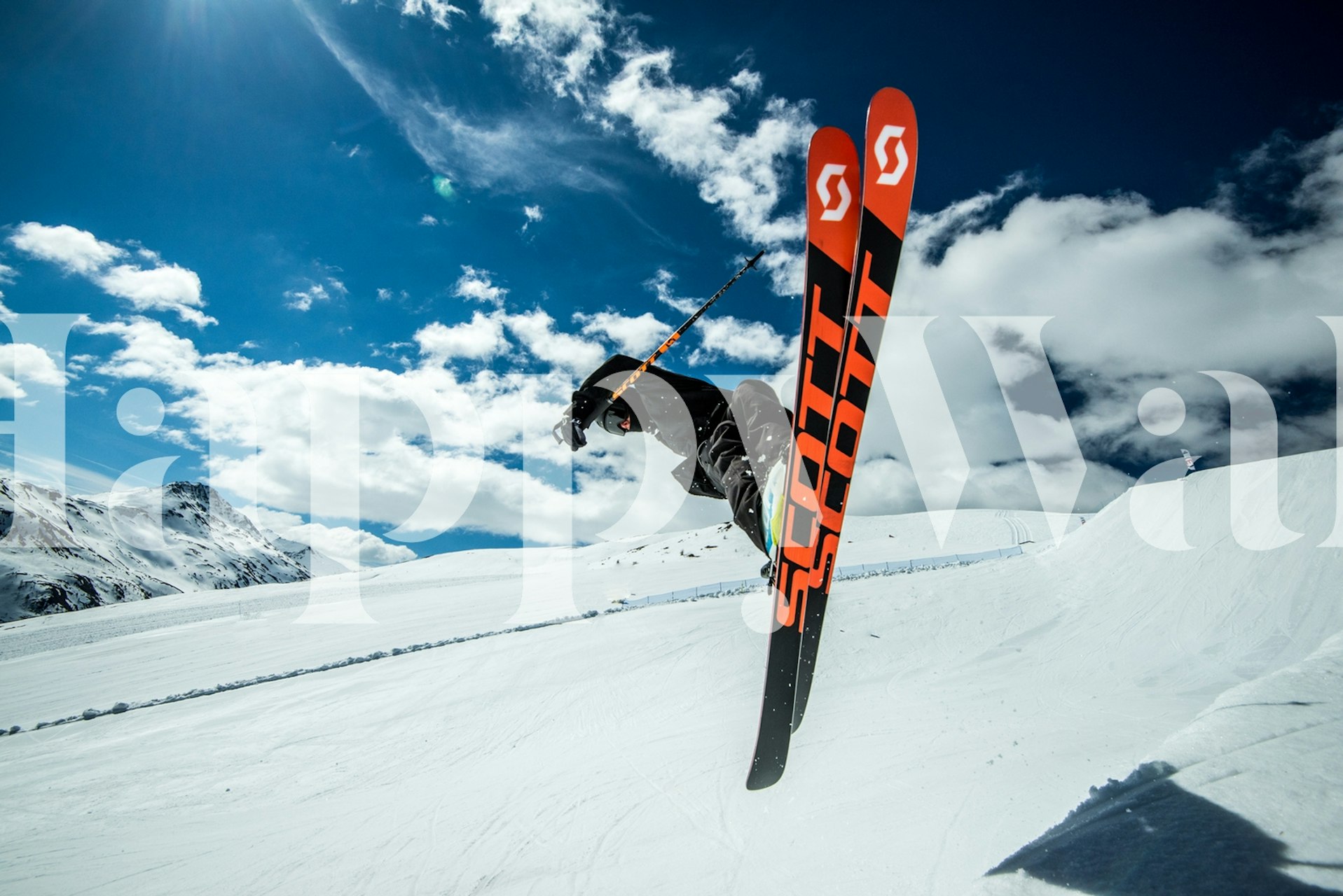 Skier in mid-air with vibrant ski gear against a snowy mountain and blue sky landscape.